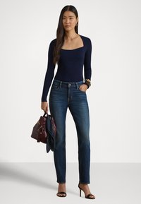 Nő tengerészkék, négyzet alakú nyakú hosszú ujjú felsőben, kék farmerral, fekete magassarkúval, kezében bordó kézitáskával és sállal, egyszínű háttér előtt áll.