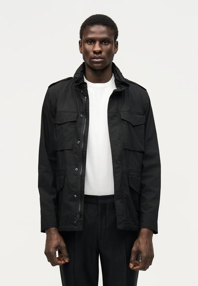Homme portant une veste utilitaire noire par-dessus une chemise blanche et un pantalon noir, debout face à un fond clair uni.