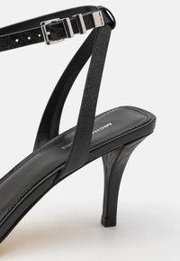 Sandale noire à talons hauts avec une tige en cuir texturé, des fines lanières, une quincaillerie en métal argenté et un design à bout carré.