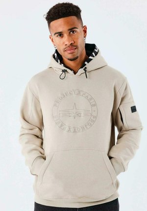 Sudadera beige con un logo redondo texturizado, capucha con cordón y bolsillo tipo canguro en la parte frontal. Presenta un cuello interno con patrón y un parche en la manga.