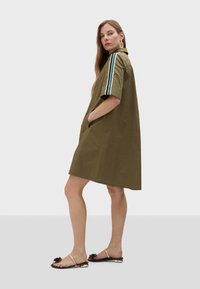 Olive green dress with short sleeves, collared neckline, side pockets, and white and green striped accents on sleeves. Worn with flat sandals.