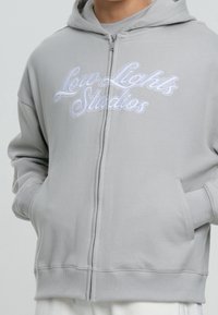 Hellgrauer Kapuzenpullover mit Reißverschluss und weiß gesticktem Text "Low Lights Studios", getragen von einer Person mit in den Taschen steckenden Händen.