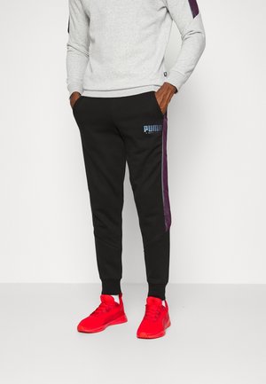 Schwarze Jogginghose mit lila Seitenstreifen, versehen mit einem blauen Puma-Logo. Kombiniert mit roten Sportschuhen und einem grauen Sweatshirt.