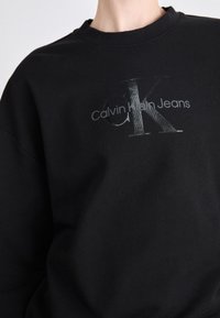 Svart hettegenser med rund hals, med en teksturert sølvlogo "CK" og "Calvin Klein Jeans" i en moderne skrifttype. Myk stofffinish.