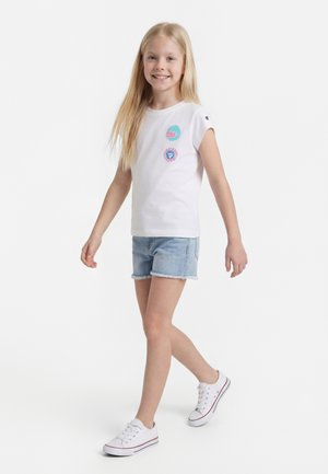 Usmievavé dievča kráča, oblečené v bielom tielku bez rukávov s farebnými záplatami, svetlé rifľové šortky a biele tenisky.