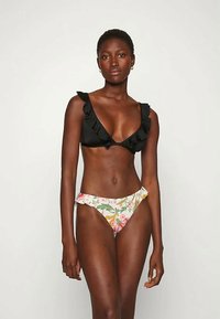 Mulher vestindo um top de biquíni preto com babados e um fundo de biquíni com estampado floral, de pé contra um fundo branco liso.