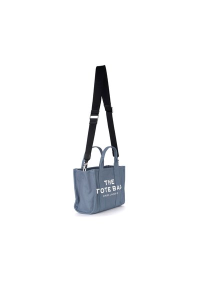 Sac en toile bleu, avec des poignées supérieures doubles et une longue sangle réglable. Un texte blanc en gros caractères indique "LE SAC TOTE". Design minimaliste.