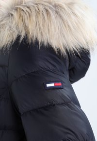 Padded black jacket with a faux fur hood. Features a logo patch with red, white, and blue accents on the left sleeve.