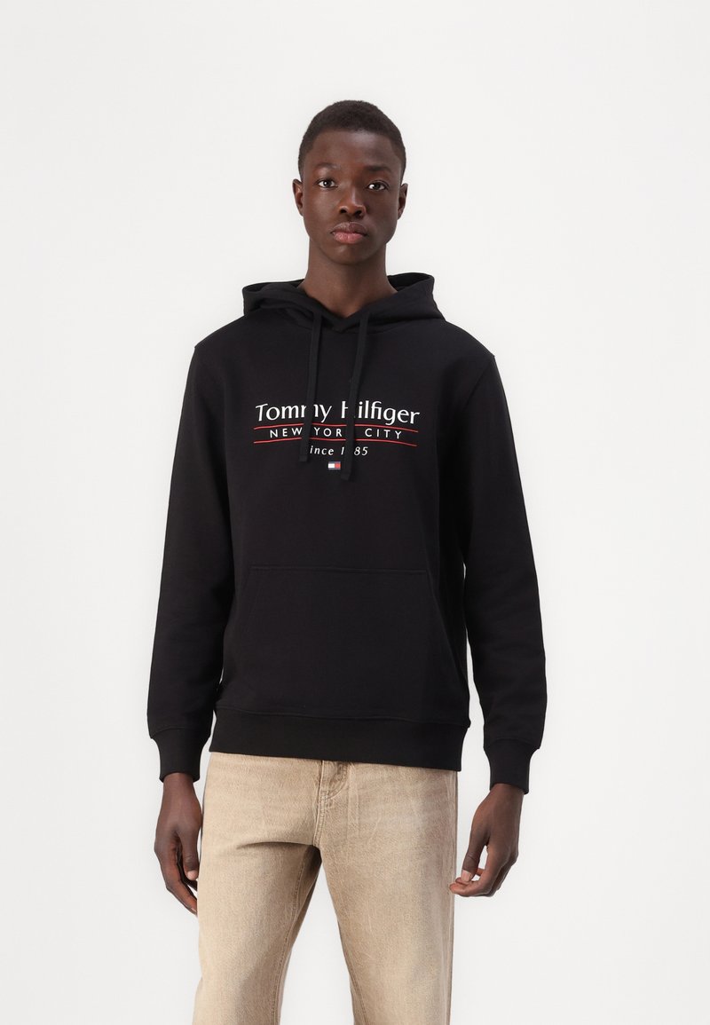 Schwarzer Hoodie aus Baumwolle mit einer Fronttasche und Kapuze mit Zugband. Prominent angezeigtes weißes "Tommy Hilfiger"-Logo mit roten und weißen Akzenten.