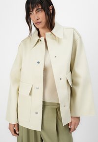 Cream-colored jacket made of thick fabric, featuring a pointed collar, buttoned front, and two patch pockets. Smooth texture with silver hardware.