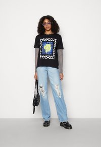 Camiseta preta com borda quadriculada e gráfico de sorriso amarelo; sobreposta com mangas longas de malha preta. Usada com jeans claros e desgastados e botas pretas.
