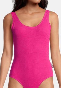 Femme portant un maillot de bain sans manches texturé rose vif avec encolure arrondie sur fond uni.