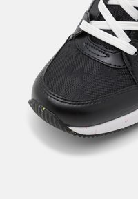 Gros plan sur la pointe d'une basket noire avec un mesh texturé, des lacets blancs, une bordure en cuir et une semelle blanche tachetée sur fond clair.