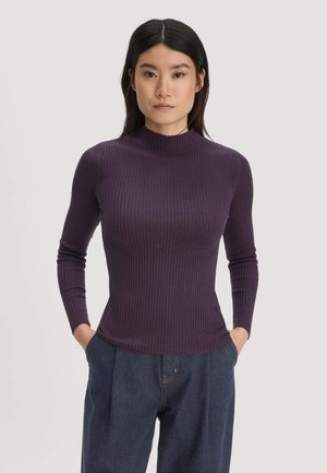 Femme aux cheveux noirs raides portant un haut à manches longues côtelé violet foncé et un pantalon bleu foncé, les mains dans les poches, face à l'avant.