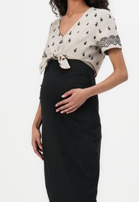 Vestido de maternidad con falda ajustada negra y parte superior beige que presenta un lazo y estampados geométricos negros. Textura suave y diseño de escote en v.
