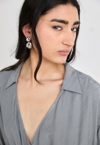 Chemise grise à col avec un décolleté en V et des rayures diagonales subtils. Grandes boucles d'oreilles rondes métalliques avec une surface réfléchissante.