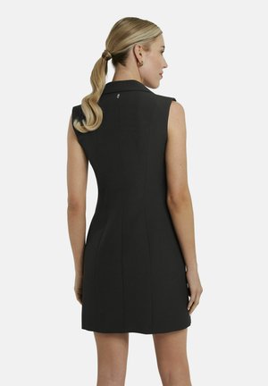 Femme avec queue de cheval blonde portant une robe noire sans manches et cintrée, vue de dos sur un fond blanc uni.