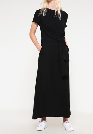 Kobieta w czarnej, krótkorękawowej sukience maxi z wiązaniem z boku, z rękami w kieszeniach, w białych trampkach na jednolitym tle.