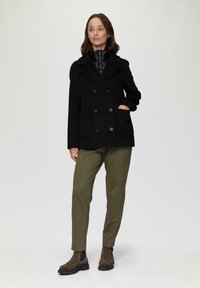 Schwarzer Peacoat mit doppelt geknöpften Knöpfen, seitlichen Taschen und Spitzkragen, kombiniert mit olivgrünen Hosen und braunen Knöchelstiefeln.