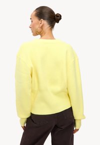 Gele oversized trui met ribbeltextuur en verlaagde schouders, gecombineerd met donkerbruine broek. Bevat een eenvoudig ronde halsontwerp.