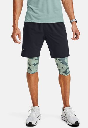 Mężczyzna ubrany w czarne krótkie spodenki sportowe na zielonych, maskujących legginsach kompresyjnych oraz białe buty do biegania, stojący z rozluźnionymi ramionami.