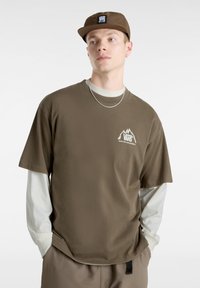 Bruine t-shirt met korte mouwen, voorzien van een bergafbeelding en het "VANS" logo, gelaagd over een licht overhemd met lange mouwen, gecombineerd met een bijpassende pet.