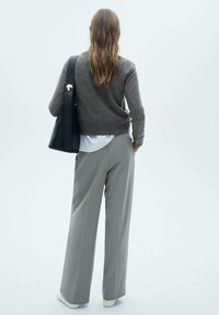 Grauer Wollpullover, hellblaues Hemd darunter, maßgeschneiderte graue Hose, schwarze Tote-Bag mit goldener Hardware. Einfarbiger Hintergrund.