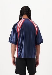 Námořnická modrá sportovní tričko s krátkým rukávem, se strapci v červené a bílé barvě po stranách a hladkým, lesklým povrchem. Pohled zezadu ukazuje kontrastní detaily.
