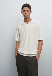 Světle béžové texturované polo triko s límečkem a krátkými rukávy, v kombinaci s tmavými kalhotami. Model stojí o proti jednobarevnému pozadí.