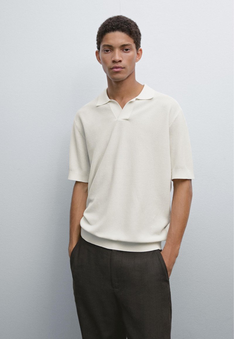 Světle béžové texturované polo triko s límečkem a krátkými rukávy, v kombinaci s tmavými kalhotami. Model stojí o proti jednobarevnému pozadí.