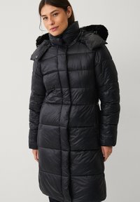 Sort polstret vinterjakke med quiltet design, høj krave og pelsforet hætte. Den har lange ærmer og lige snit, lavet af syntetisk materiale.
