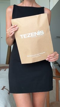 Hnědý papírový sáček s bílým potiskem, na kterém je nápis "TEZENIS spodní prádlo" a symbol recyklace na dně. Sá�ček drží osoba v černých šatech.