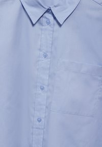 Camisa de color azul claro con cuello, hecha de una tela suave, que cuenta con botones en la parte delantera y un bolsillo en el pecho. Diseño simple sin patrones.