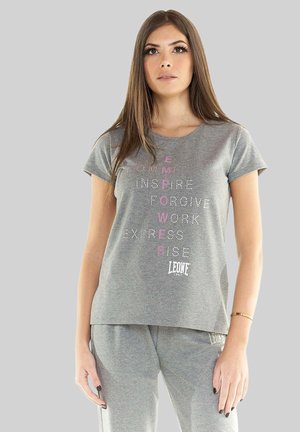 Jeune femme debout portant une tenue grise avec des mots positifs et "EMPOWER" en lettres roses verticalement sur le t-shirt.
