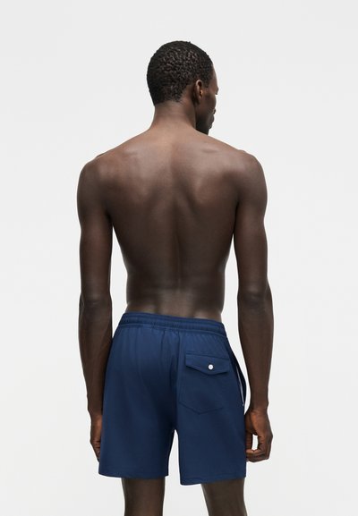 Άνδρας με σκουρόχρωμο δέρμα που φοράει σκούρα μπλε μαγιό, εμφανιζόμενος από πίσω σε απλό λευκό φόντο.