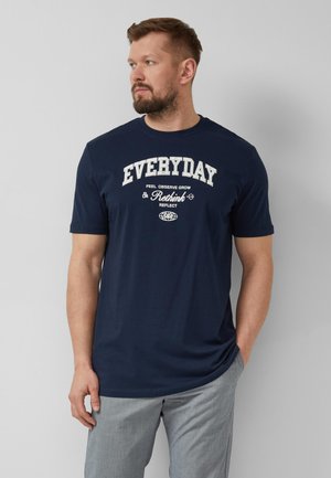 Mann mit Bart, der ein dunkelblaues T-Shirt mit weißem "EVERYDAY" Schriftzug und hellgraue Hose trägt, vor einem einfachen hellgrauen Hintergrund stehend.