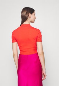 Top corto rossa a costine con maniche corte e collo alto, con dettagli bianchi sulle spalle. Sfondo grigio uniforme.