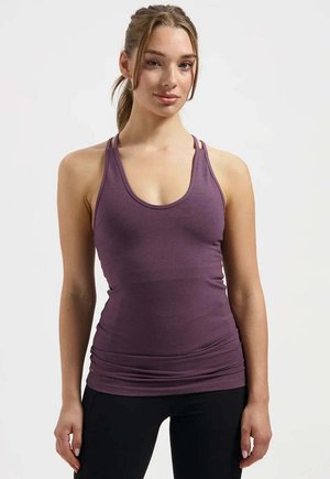 Jeune femme aux cheveux châtain clair attachés, portant un débardeur violet ajusté et des leggings noirs, debout devant un fond blanc uni.