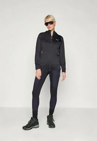 Person som har på seg svart zip-up jakke, svarte leggings med lommer, svarte tursko og mørke solbriller, står mot en enkel lys bakgrunn.