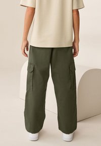 Olívovo zelené cargo nohavice s veľkými bočnými vreckami, kombinované s svetlo béžovou textúrovanou košeľou. Oblečenie dopĺňajú biele tenisky.