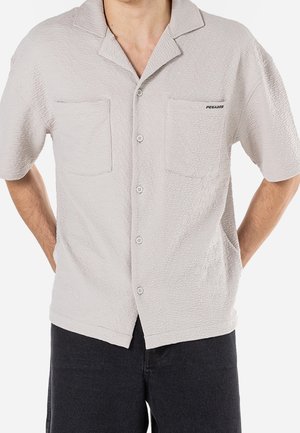Beige Kurzarm-Hemd mit Knopfleiste aus strukturiertem Stoff, zwei Brusttaschen und einem Reverskragen. Markennamen auf der Tasche.