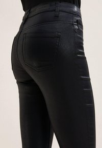 Schwarze, hochtaillierte Denim-Jeans mit glänzender Textur. Merkmale sind Gesäßtaschen und eine figurbetonte Beinsilhouette. Minimalistisches Design.