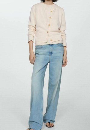 Vrouw draagt een crèmekleurig vest met knopen, gecombineerd met lichtblauwe wijde jeans en zwarte sandalen met open tenen, staande tegen een effen achtergrond.