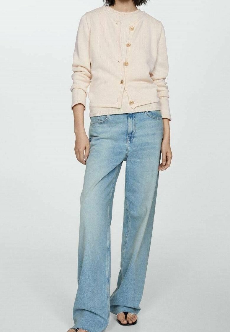 Femme portant un cardigan crème boutonné associé à un jean large bleu clair et des sandales noires à bout ouvert, debout devant un fond uni.