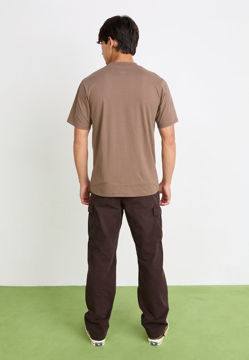 T-shirt en coton marron à manches courtes, associé à un pantalon cargo foncé avec poches latérales, se tenant sur un sol en moquette verte.