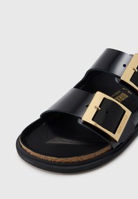 Μαύρο σανδάλι από δέρμα με γυαλιστερή επένδυση, που διαθέτει δύο φαρδιές λουριά με χρυσά μεταλλικά αγκράφες, πάτο από φελλό και υφή στη σόλα από καουτσούκ.