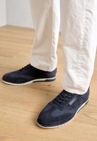 Marina mockasiner i mocka med texturerade detaljer och en tunn vit sula, tillsammans med ljusa, avslappnade byxor på ett trägolv.