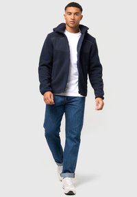 Tumedast fleece-jakk, millel on lukuga esiosa, kapuuts ja kontrastse rinnaosa, koos siniste teksade ja heledad sneaksiga. Lõdva istuvusega.