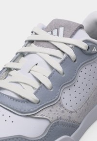 Zbliżenie na biało-szary sneaker z perforowaną i teksturowaną skórą, białymi sznurowadłami oraz logo na języku i boku.