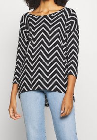 Musta ja harmaa chevron-kuvioinen yläosa, jossa on leveä kaula-aukko ja 3/4-hihat, yhdistettynä vaaleansinisiin farkkuihin. Materiaalin vaikuttaa olevan pehmeä ja kevyt.